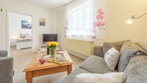 A modern living room with a sofa, a coffee table with books and flowers, a TV, and a window with floral decorations. - Apartment in Prerow up to 2 p. + child up to 6 y., quiet, close to the beach, parking lot, WLAN (Prerow)