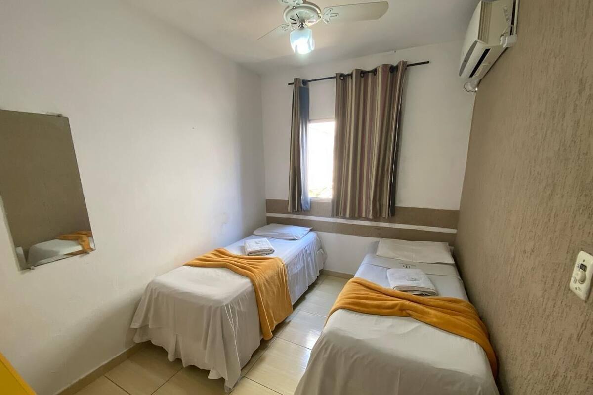 Una habitación pequeña y limpia con dos camas individuales, un ventilador de techo, una ventana con cortinas y un equipo de aire acondicionado.