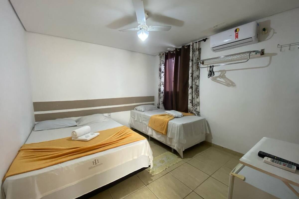 Habitación de hotel con dos camas, ventilador de techo, equipo de aire acondicionado, una mesita con un control remoto y una ventana con cortinas.