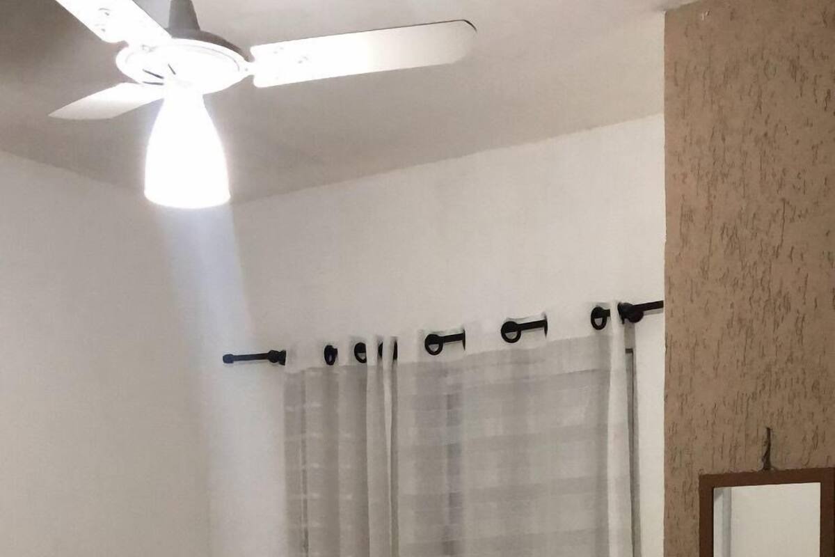 Un dormitorio con cama, ventilador de techo, ventana con cortinas y espejo en la pared.