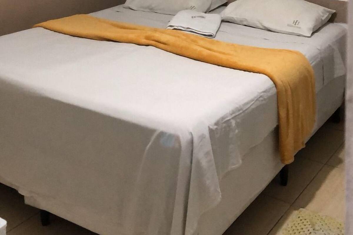 Una cama individual con un colchón blanco y una manta amarilla, un ventilador de techo y cortinas de color claro.