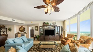 A living room with a sofa, armchairs, a coffee table, and a TV. There is a ceiling fan, a large window with a view, and a kitchen in the background. - Three Bedroom Condo with Lazy River & Gulf of Mexico Views. Complimentary Boat Slip! (Orange Beach)