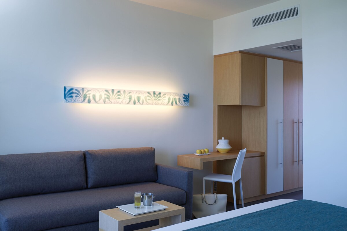 Une chambre d’hôtel moderne avec un canapé gris, une petite table en bois sur laquelle se trouve un verre d’eau et un pichet en métal, une chaise blanche et un lit recouvert d’une couverture bleue.
