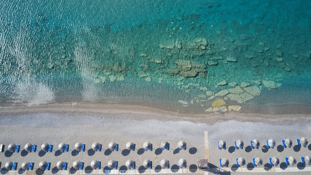 Vista aérea de una playa con agua turquesa cristalina, costa rocosa y filas de tumbonas.