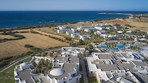 Vista aérea de un complejo turístico costero con varias edificaciones blancas, una piscina y vistas al océano.