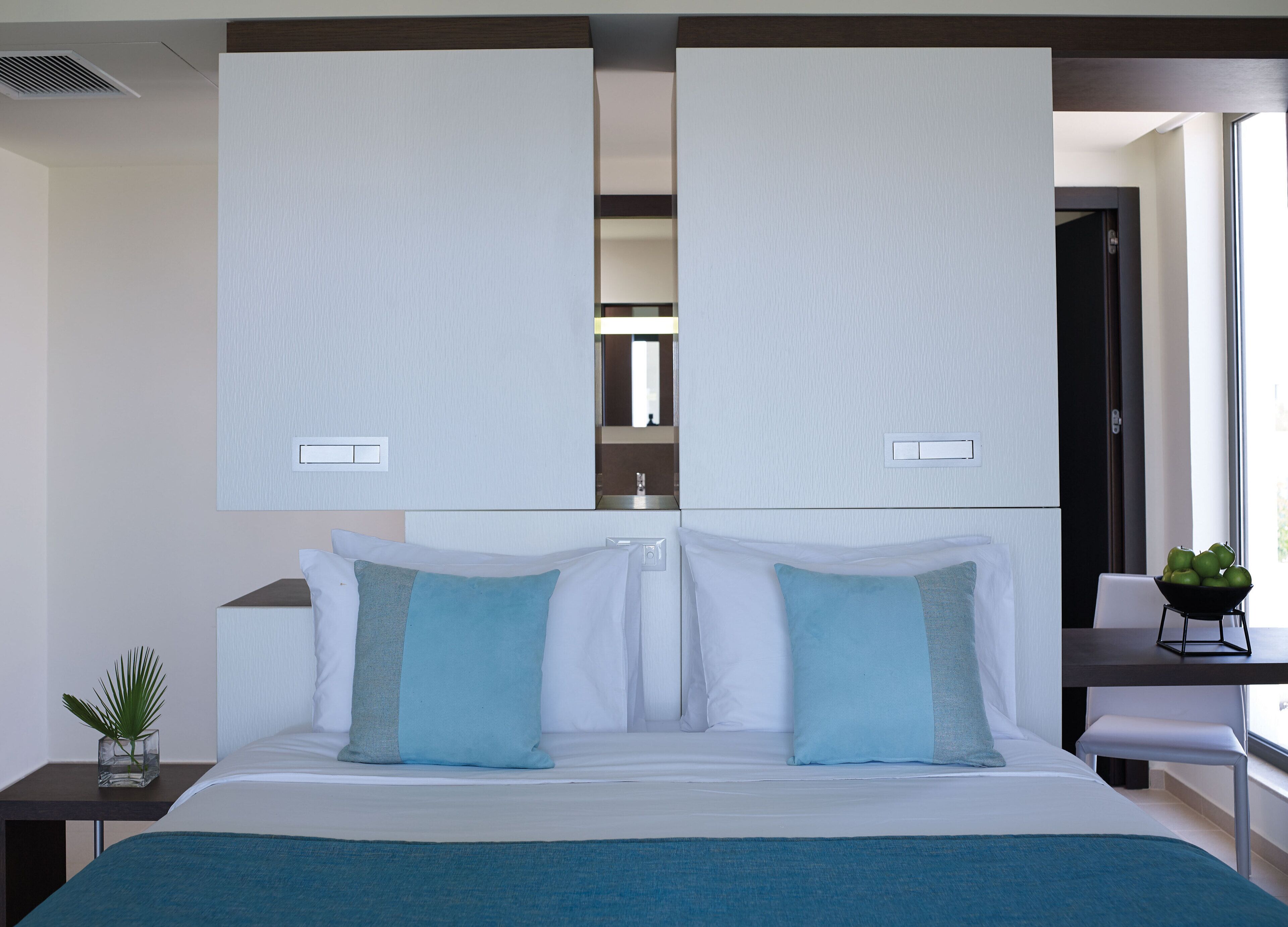 A modern hotel room with a large bed, two nightstands, a small table with a bowl of fruit, and a view of the outside through a window.