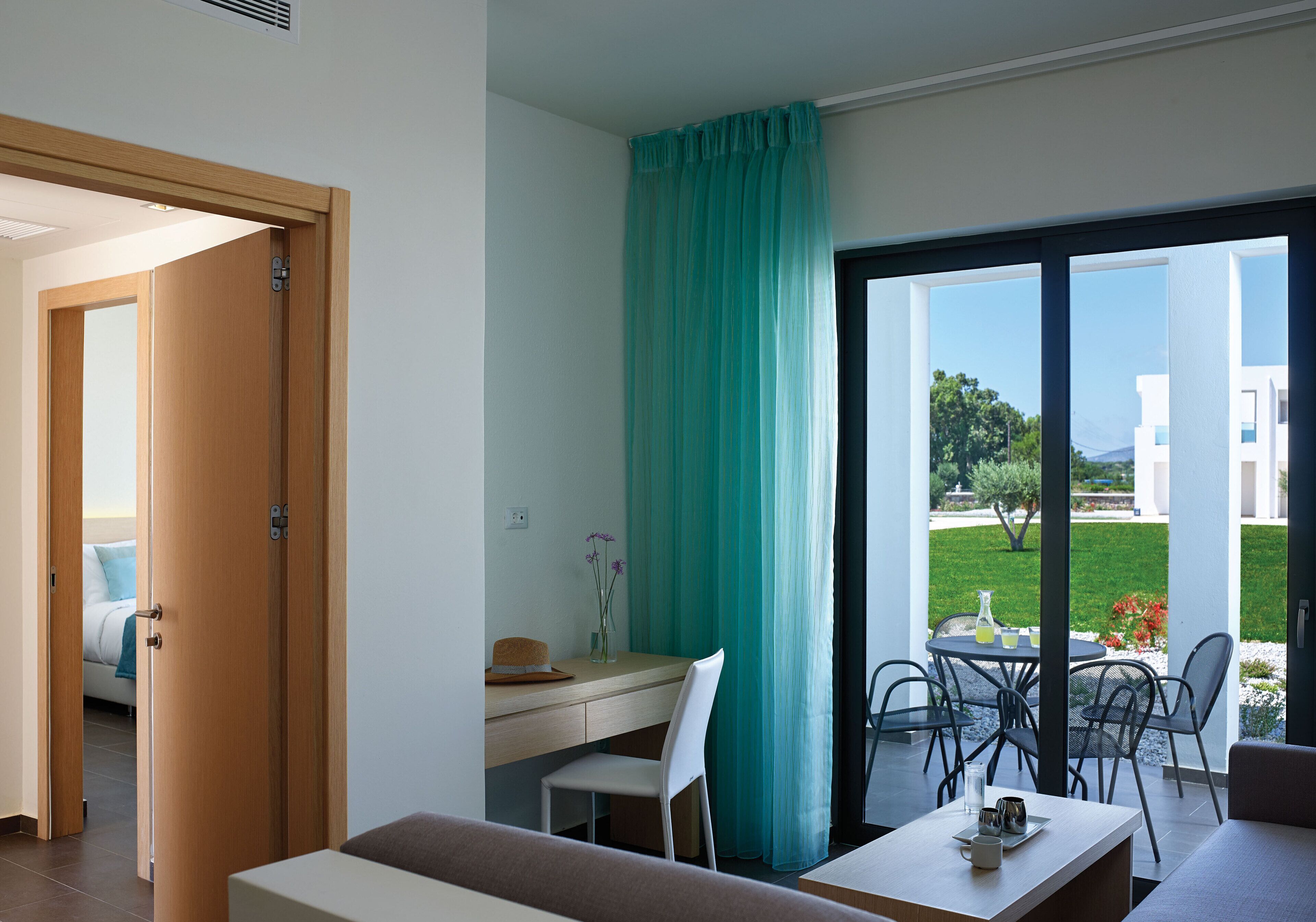 A modern room with a desk, a chair, a table with a vase, and a view of a garden and buildings outside.