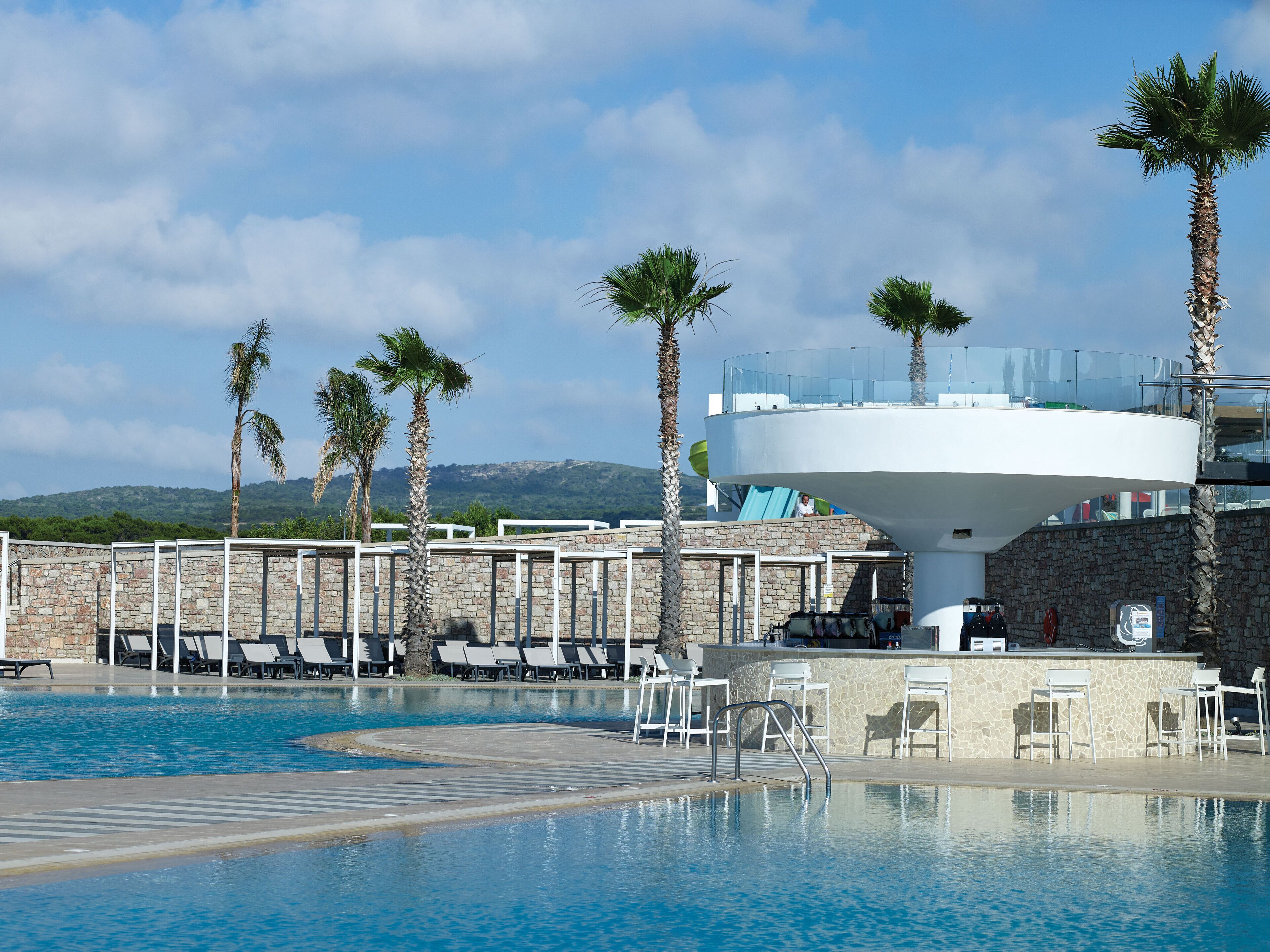 A swimming pool with lounge chairs and palm trees, surrounded by stone walls and a clear sky.
