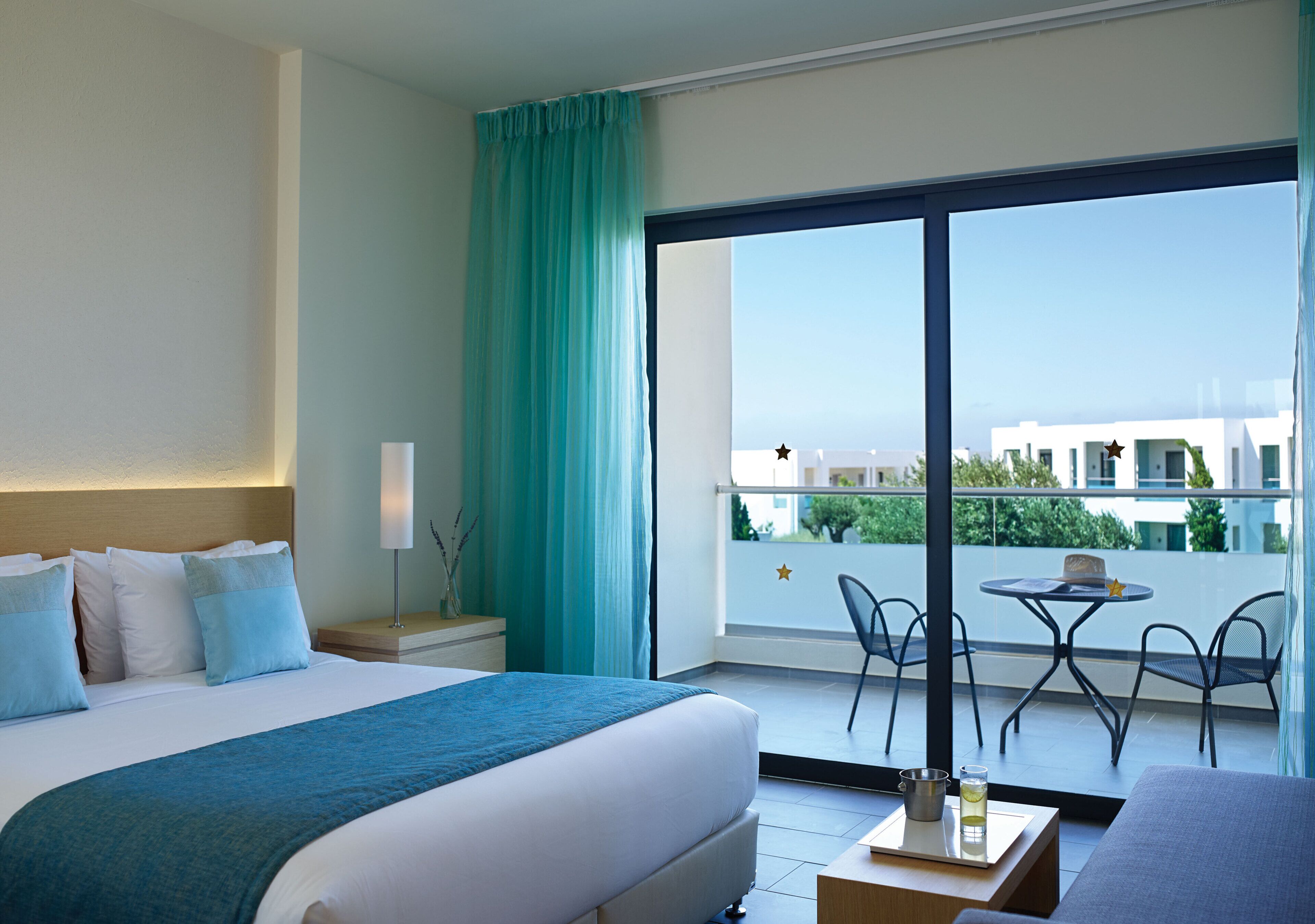 Una camera d'hotel con un letto, un balcone con sedie e un tavolo, e vista sugli edifici.