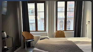 Habitación de hotel con cama, dos sillas, un radiador y una ventana con vistas a edificios.