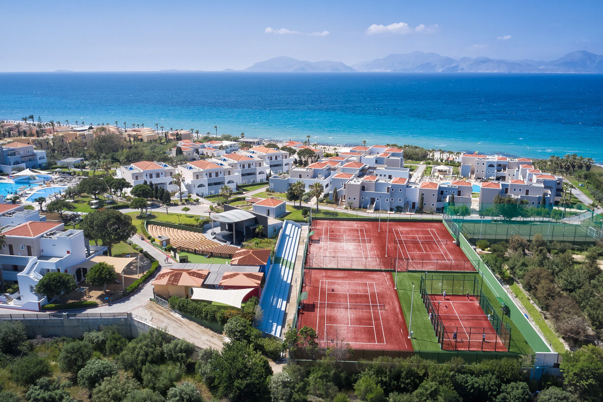 An aerial view of a coastal resort with multiple white buildings, a tennis court, and a pool area.