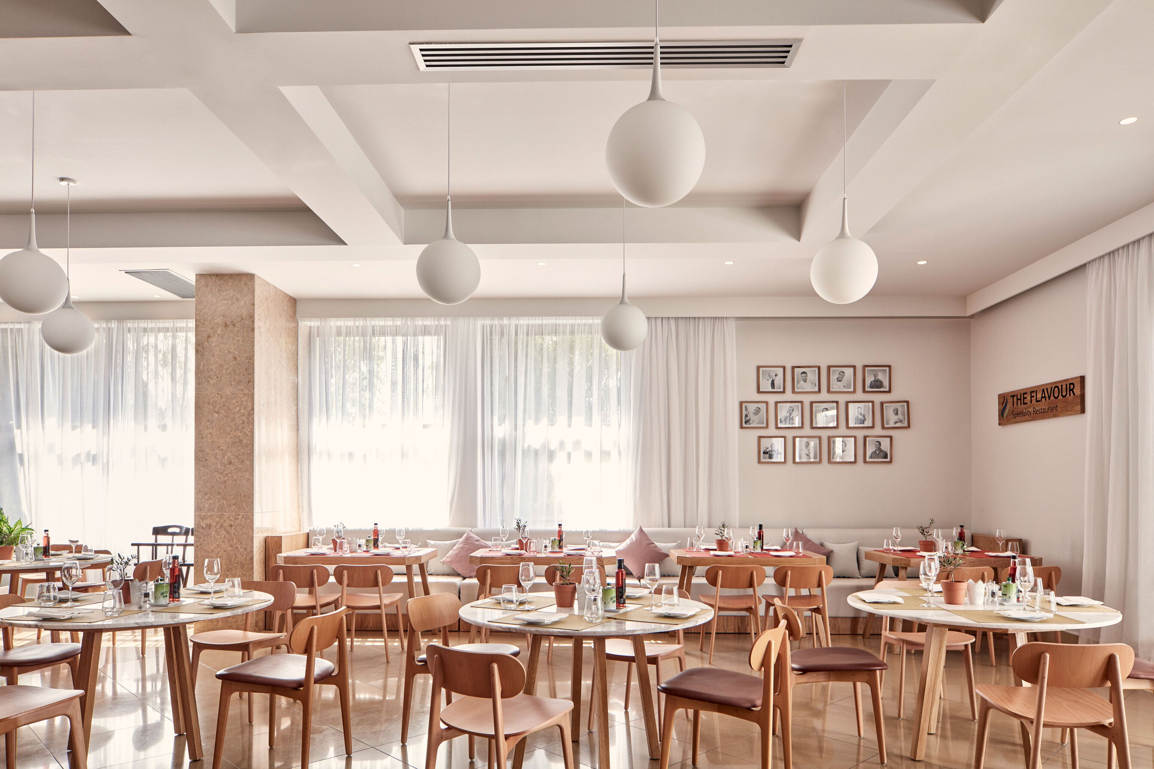 A modern dining area with round tables and chairs, pendant lighting, and a large window with sheer curtains.