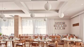 A modern dining area with round tables and chairs, pendant lighting, and a large window with sheer curtains.