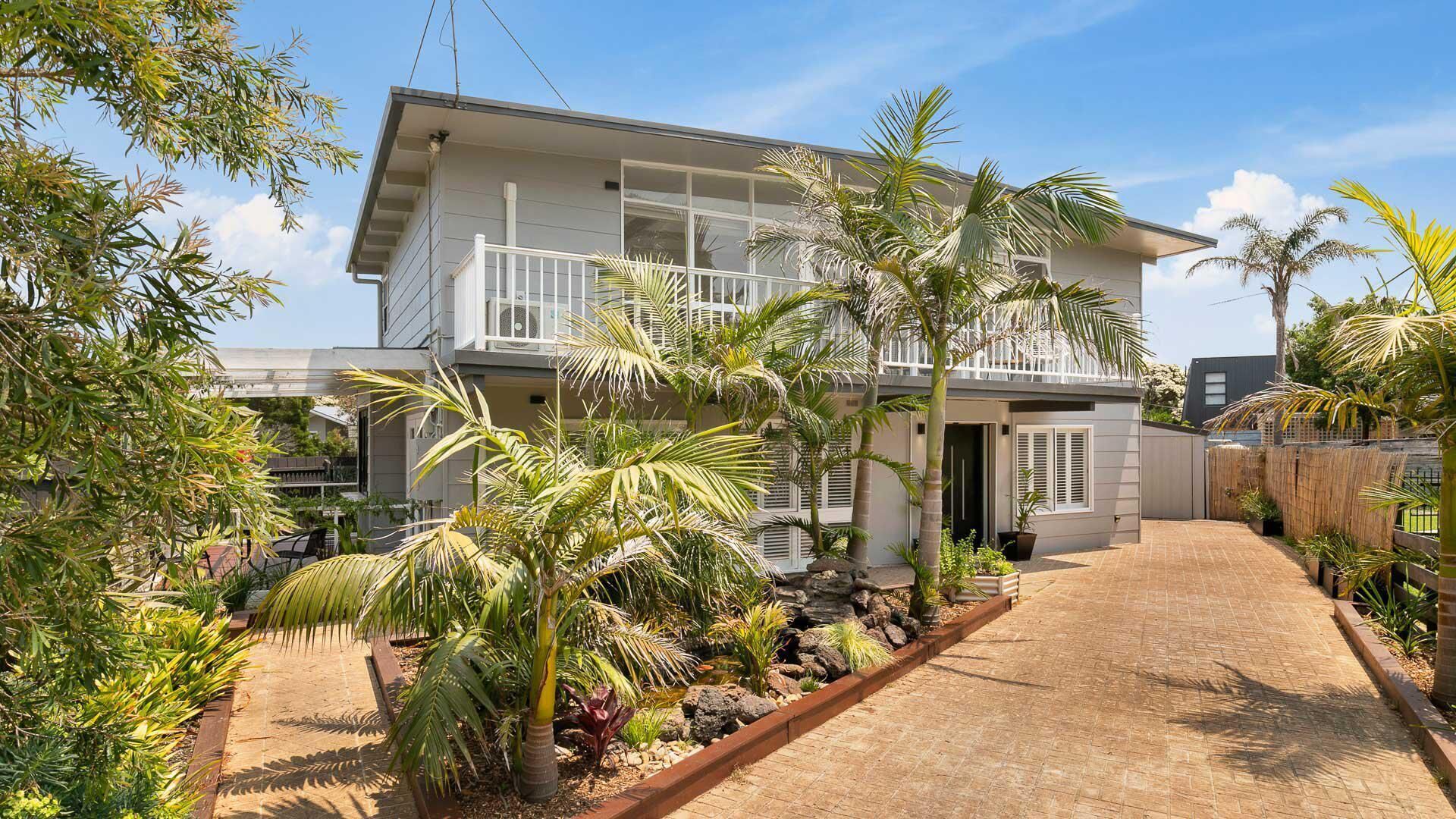 A two-story house with a balcony, surrounded by tropical plants and a paved driveway.