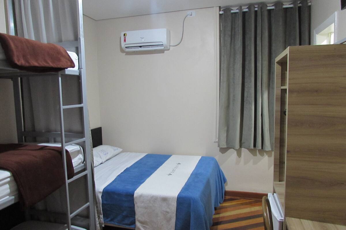 Un dormitorio con literas, una cama individual con cubrecama azul y blanco a rayas, un equipo de aire acondicionado, un armario de madera y una ventana con cortinas.