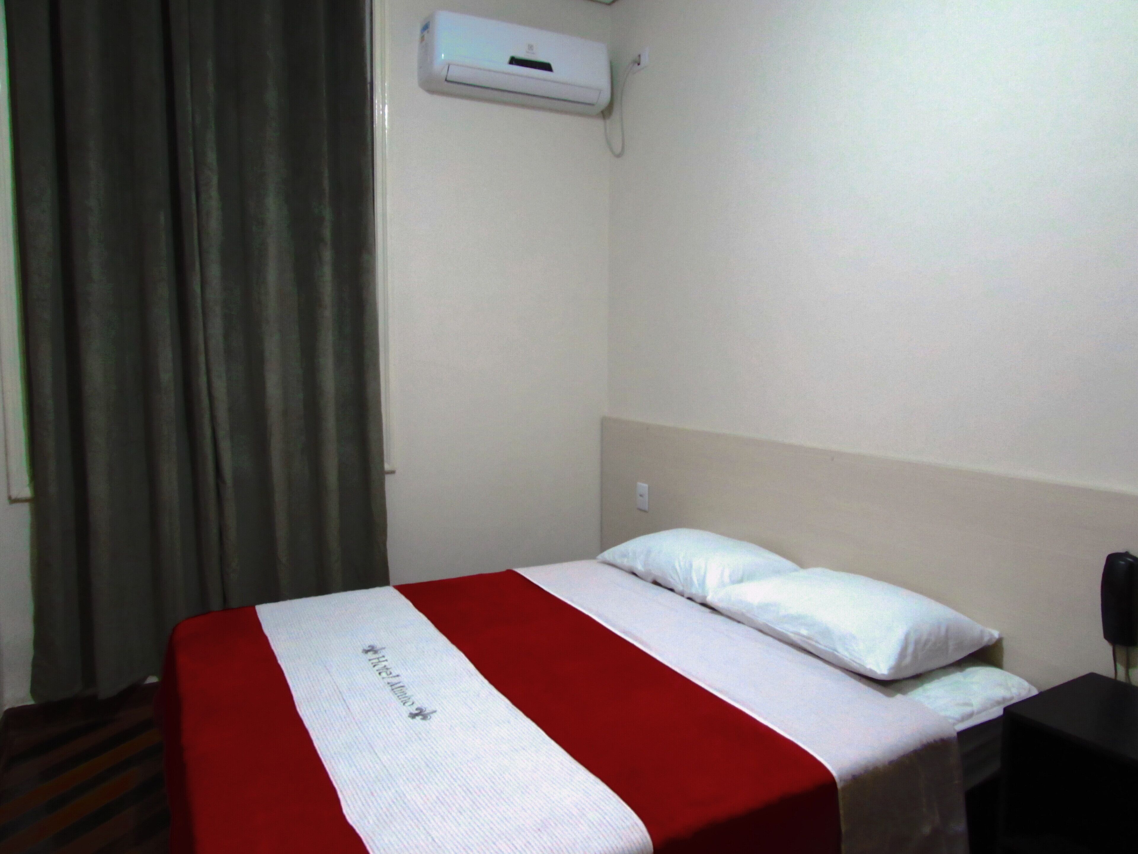A hotel room with a bed, a red and white bedspread, a white pillow, a nightstand, a black phone, and a wall-mounted air conditioning unit.