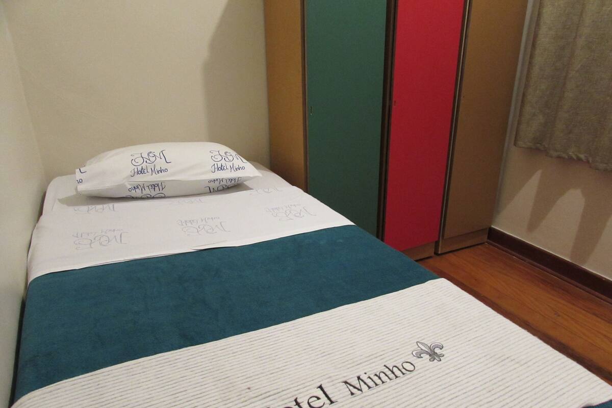 Una cama individual con colcha blanca y verde azulada, almohadas y una manta con la inscripción "Hotel Minho".