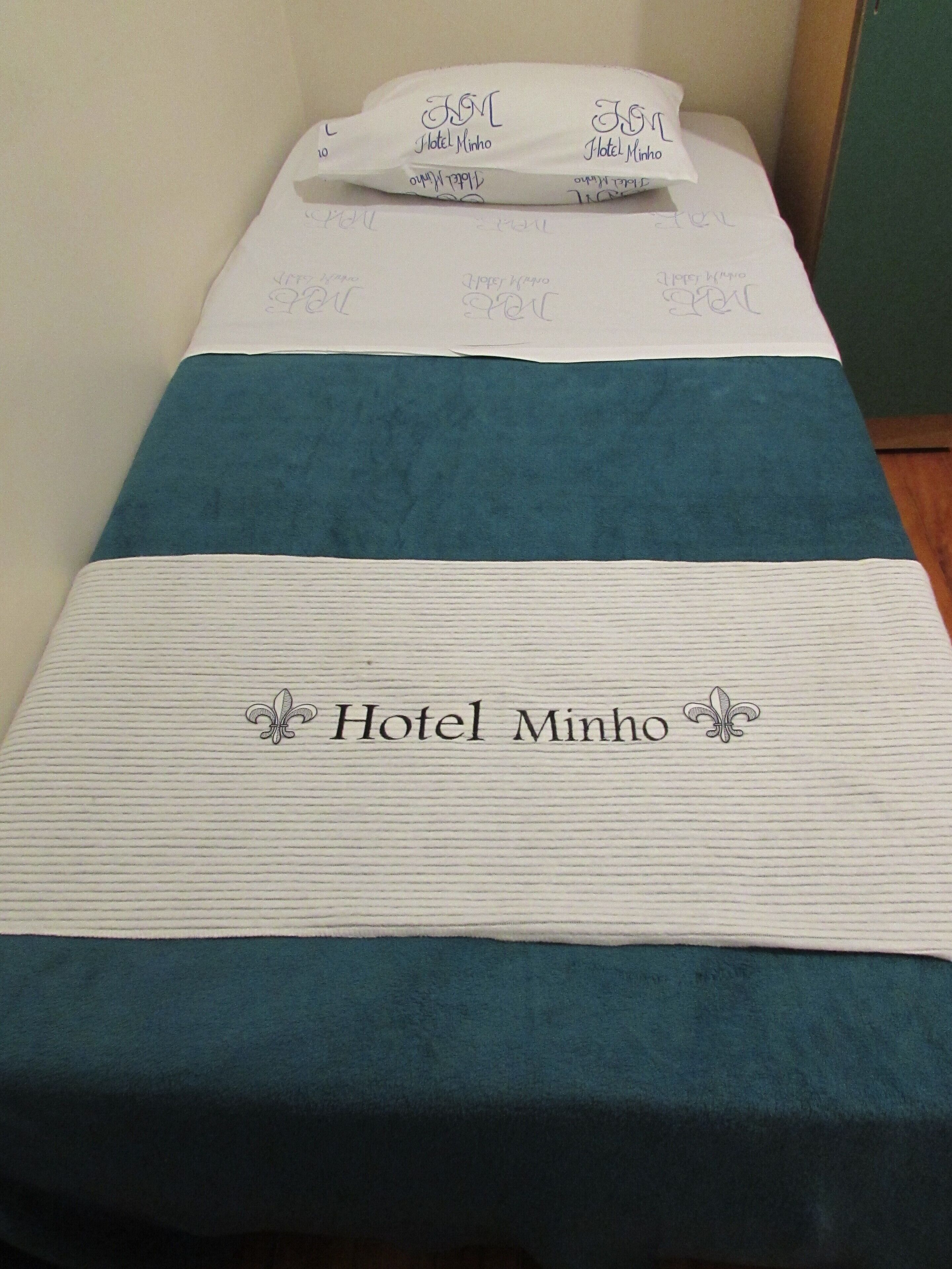 A hotel bed with a teal bedspread and a white pillow with 'Hotel Minho' embroidered on it.