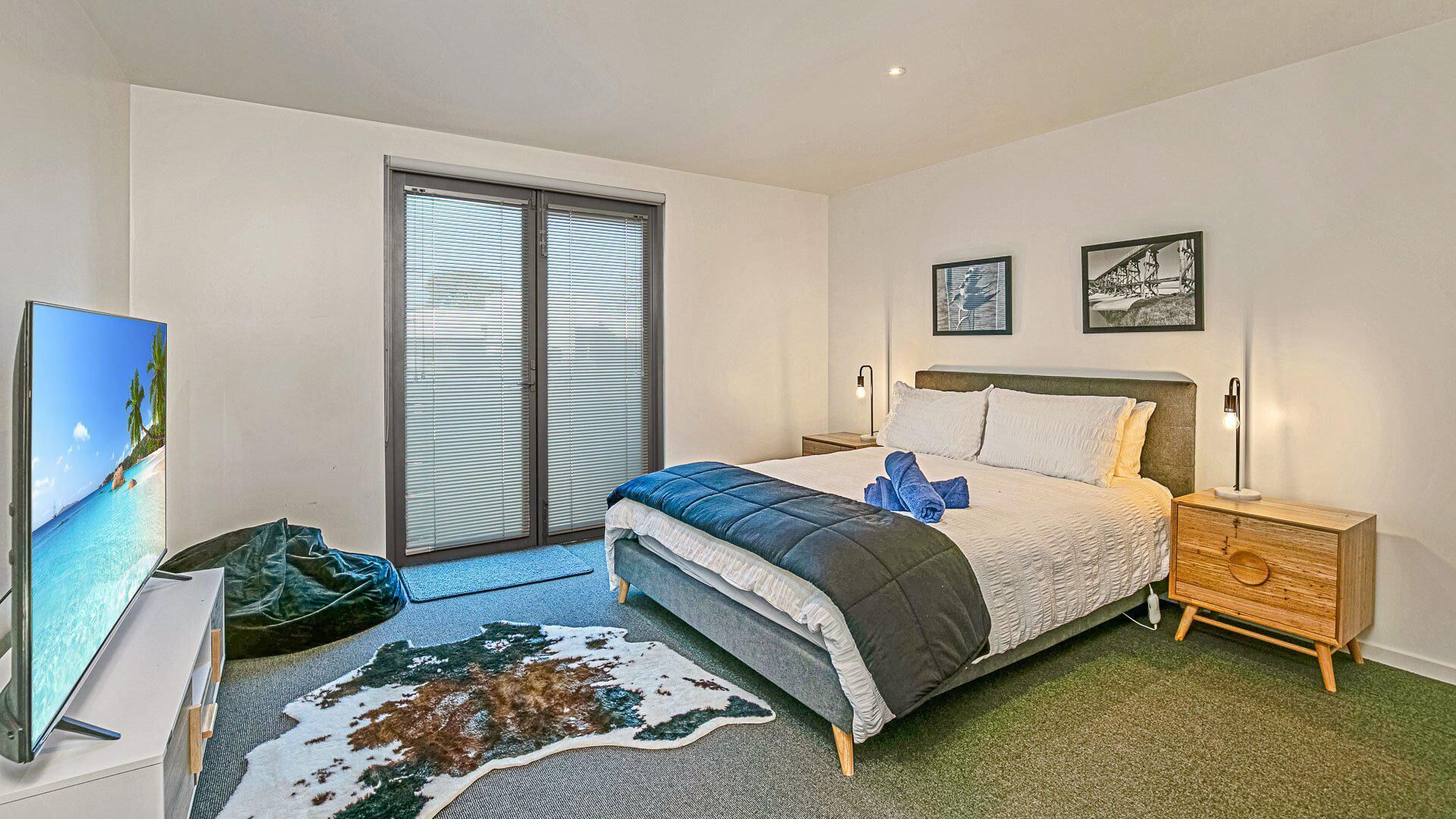 Une chambre à coucher comprenant un lit, une table de chevet, un téléviseur, une fenêtre avec des stores et deux tableaux encadrés accrochés au mur.