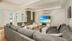 Una sala de estar moderna con un sofá gris grande, una mesa de centro y un televisor de pantalla plana mostrando una escena de playa. Hay dos ventanas con vistas a zonas verdes y un techo con iluminación empotrada.