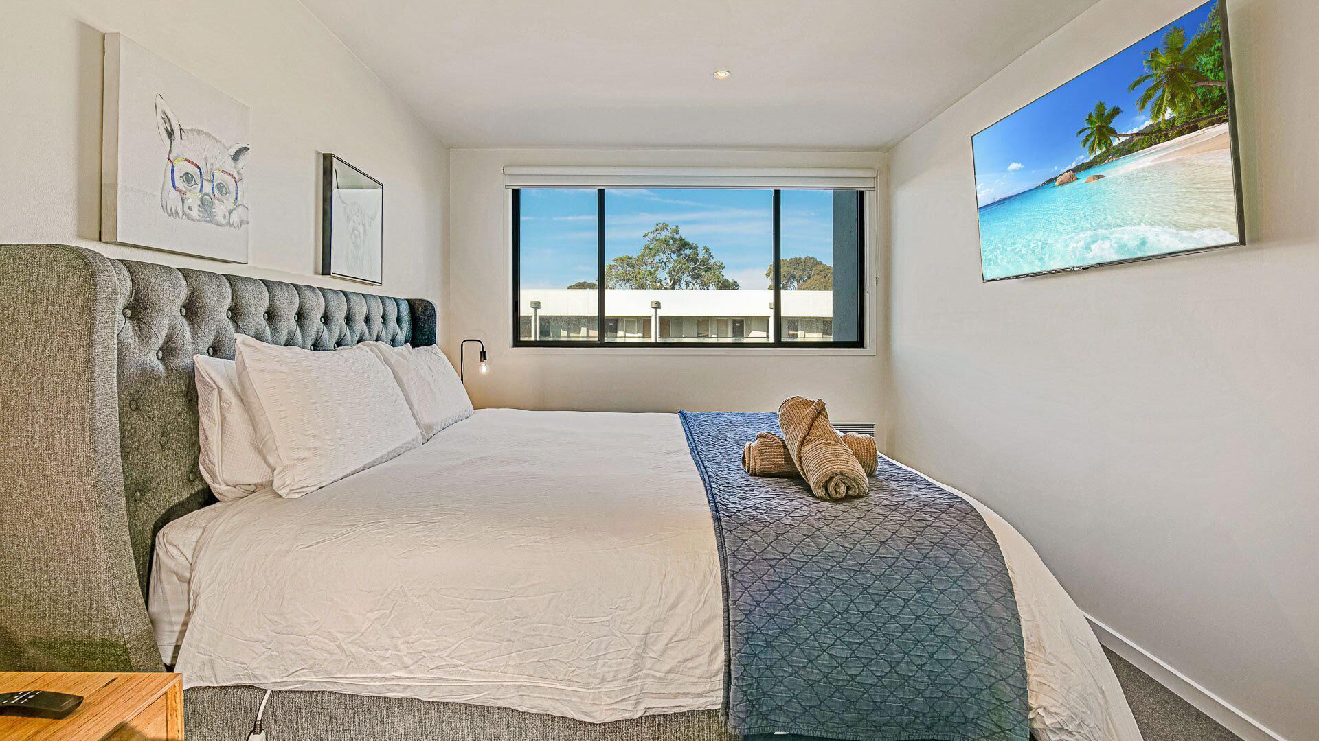 A bedroom with a large bed, a window with a view of trees, and a painting of a beach.
