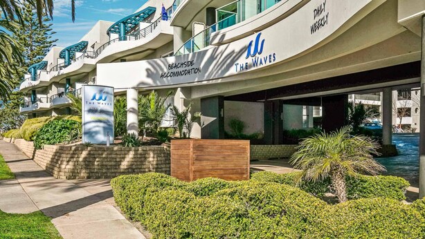 Un hotel frente a la playa llamado 'The Waves' con un diseño moderno, que cuenta con balcones y un cartel anunciando alojamiento y habitaciones disponibles en la playa.