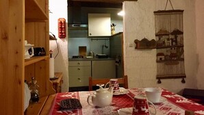 A cozy dining area with a table set for two, a wooden shelf with kitchen items, and a kitchen visible in the background. - SUITE ALPAGE (Méolans-Revel)