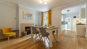 A dining area with a wooden table, four chairs, a yellow armchair, a fireplace, and a view into the kitchen.