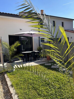 A modern outdoor patio with a dining area, surrounded by lush greenery and a white building in the background. - The banks of the Isle (Périgueux)