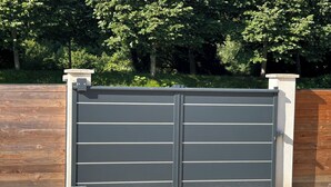 Une porte métallique fermée, de couleur grise, flanquée de deux piliers blancs, se dressant sur fond de clôture en bois et de feuillage dense.