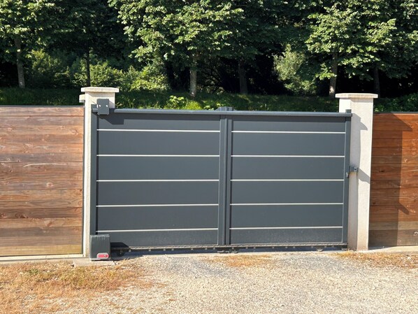 A closed metal gate with a grey finish, flanked by two white pillars, set against a backdrop of a wooden fence and dense foliage.