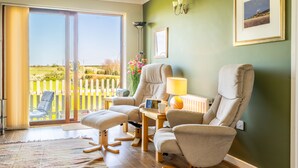 Ein Wohnzimmer mit einer großen Schiebeglastür, die auf einen Balkon führt, einem Sofa, einem Tisch mit einer Lampe und einem gerahmten Bild an der Wand.