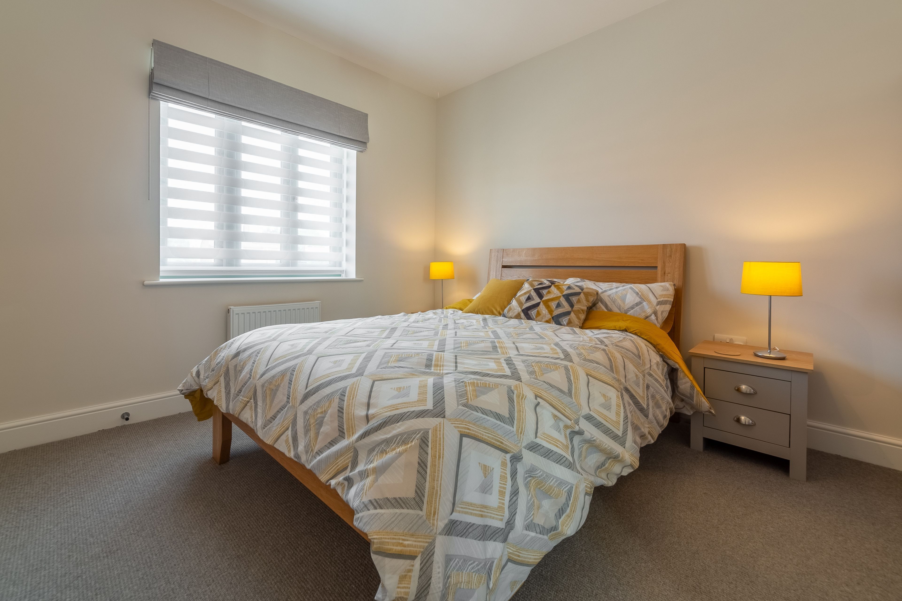 A bedroom with a bed, two bedside tables with lamps, a window with blinds, and a radiator.