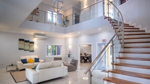 A spacious living area with a white sofa set, a dining table, and a staircase with wooden steps and glass railings. - The Sun Seeker Villa Ja 9 Bedrooms . Fully staffed with private chef & butler  (Montego Bay)