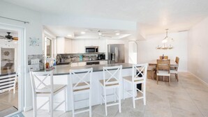 A modern kitchen with white cabinetry, a central island with bar stools, and a dining area with wooden chairs. - Island Oasis-Waterfront Pool home! 10 min walk to the beach! (Marco Island)