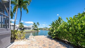 A paved walkway leading to a body of water, flanked by palm trees and a building with a glass facade. - Island Oasis-Waterfront Pool home! 10 min walk to the beach! (Marco Island)