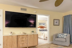 A living room with a TV, wooden cabinet, grey sofa, and a bed in the background.