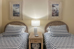 Une chambre avec deux lits, chacun agrémenté de draps à rayures, une table centrale avec une lampe, et deux tableaux encadrés accrochés au mur.