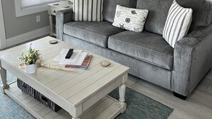 A living room with a grey sofa, striped pillows, a round wall art piece, a white coffee table, and a window with blinds. - Beachfront-Top floor, corner. Waterfront views! Smart TVs and WiFi. Sleeps 2 (Tybee Island)