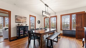 Un vaste espace dédié à la salle à manger, avec une longue table en bois, des chaises noires et un banc. On y trouve un buffet noir aux portes vitrées, une horloge au mur et de grandes fenêtres à cadres en bois.