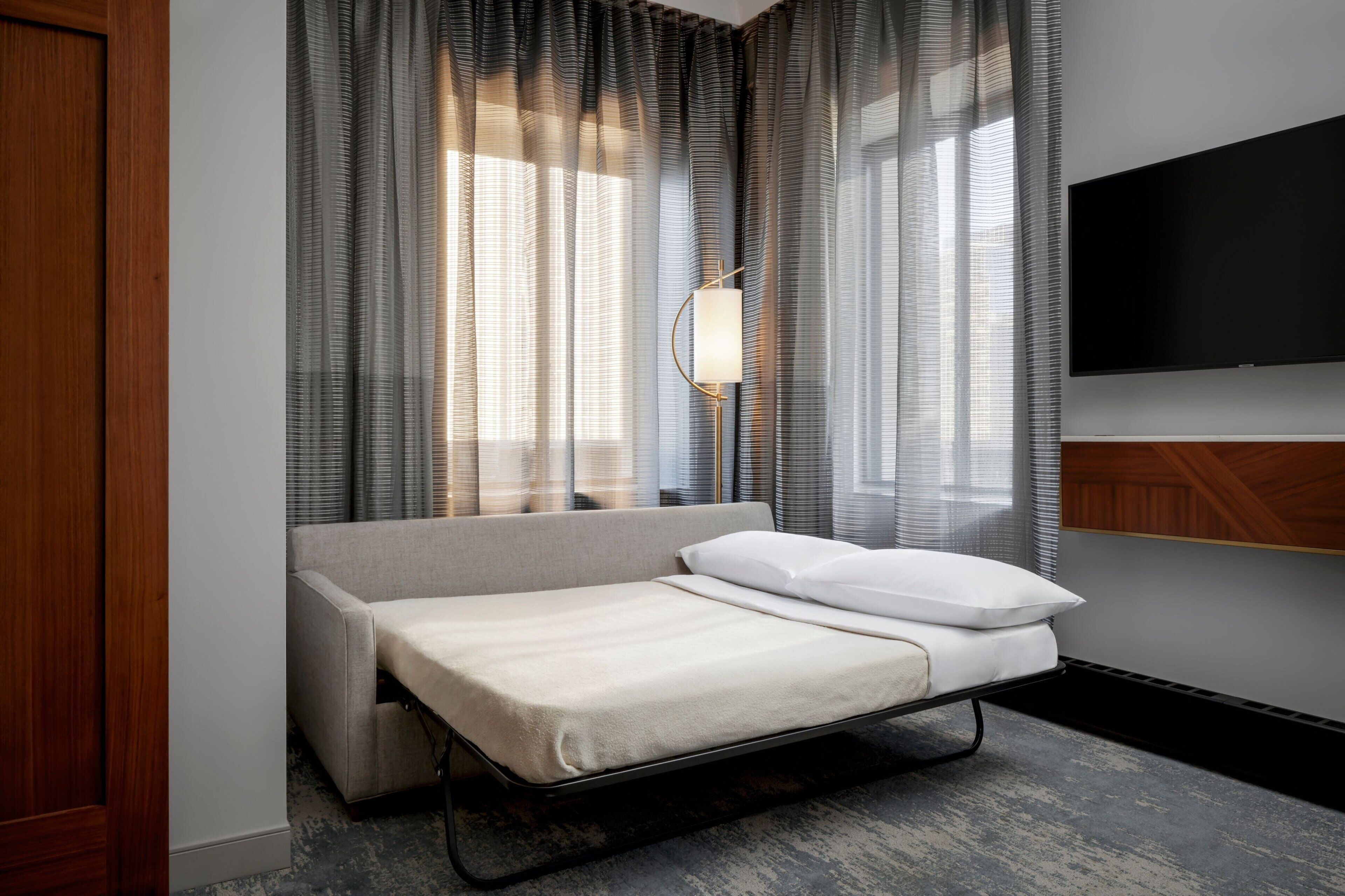 A hotel room with a bed, a TV mounted on the wall, and a window with sheer curtains.