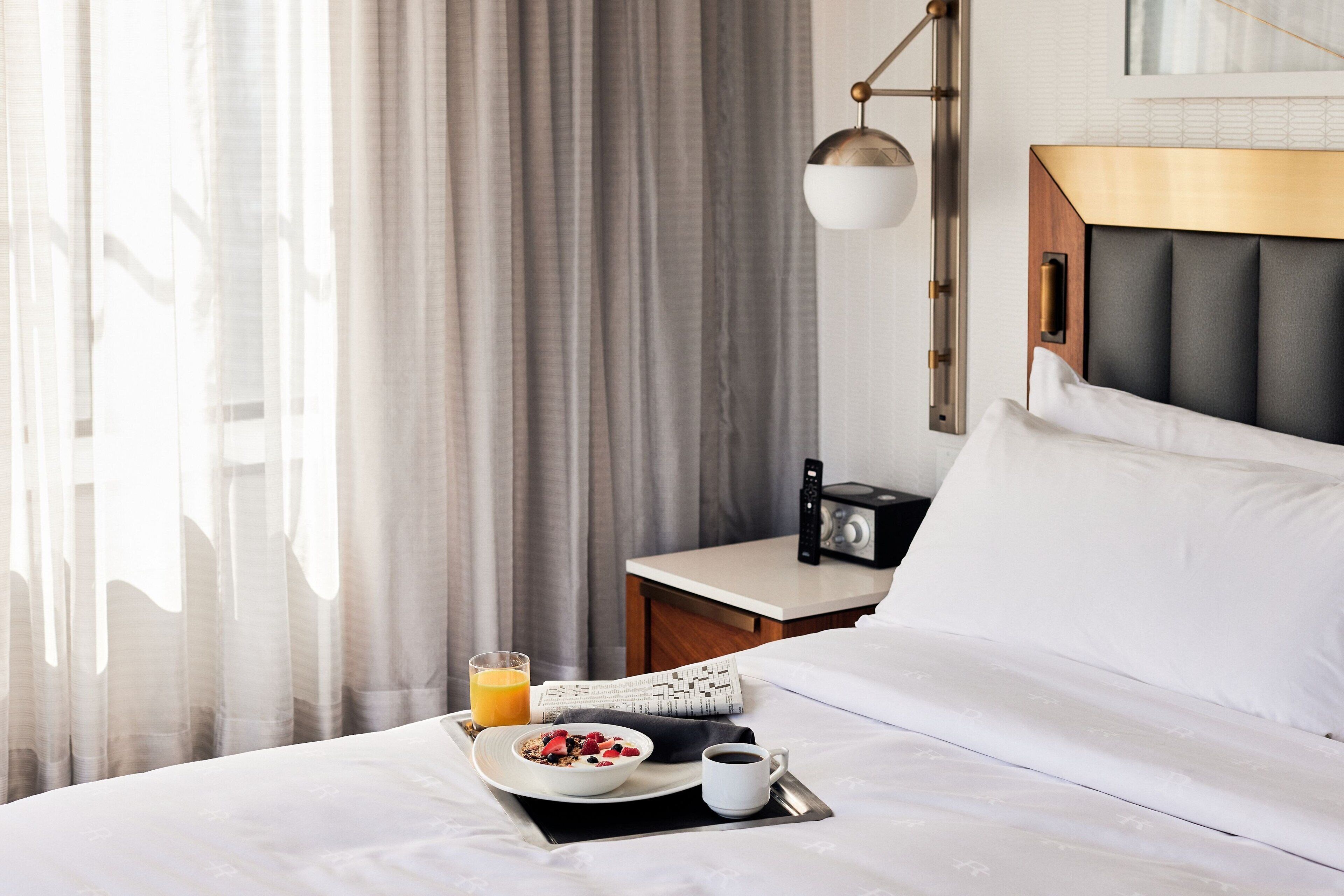 A neatly made bed with white linens, a bedside table with a radio, a tray with breakfast including cereal, fruit, coffee, and orange juice.