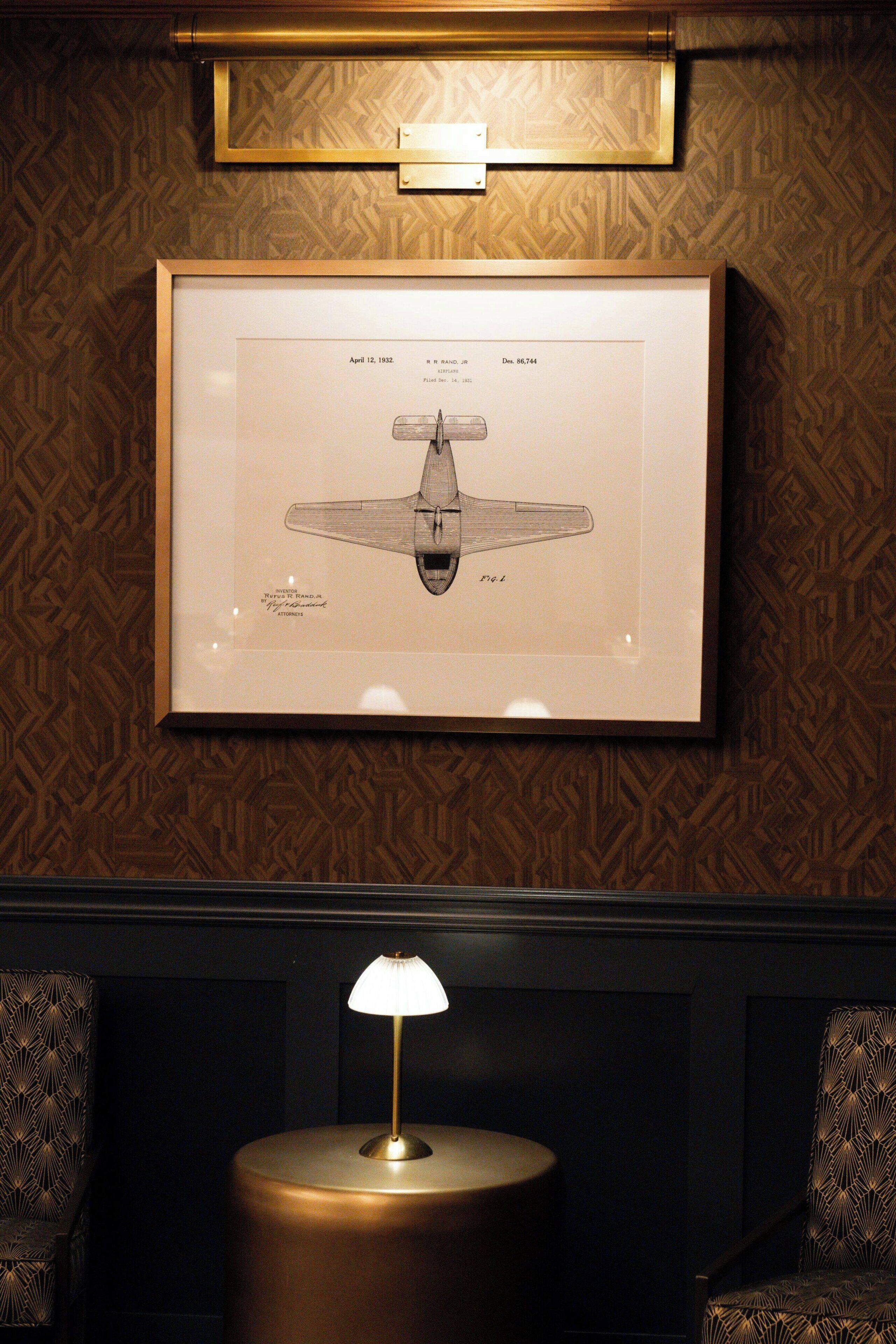 A framed airplane drawing on a patterned wall, a round table with a lamp, and patterned chairs.