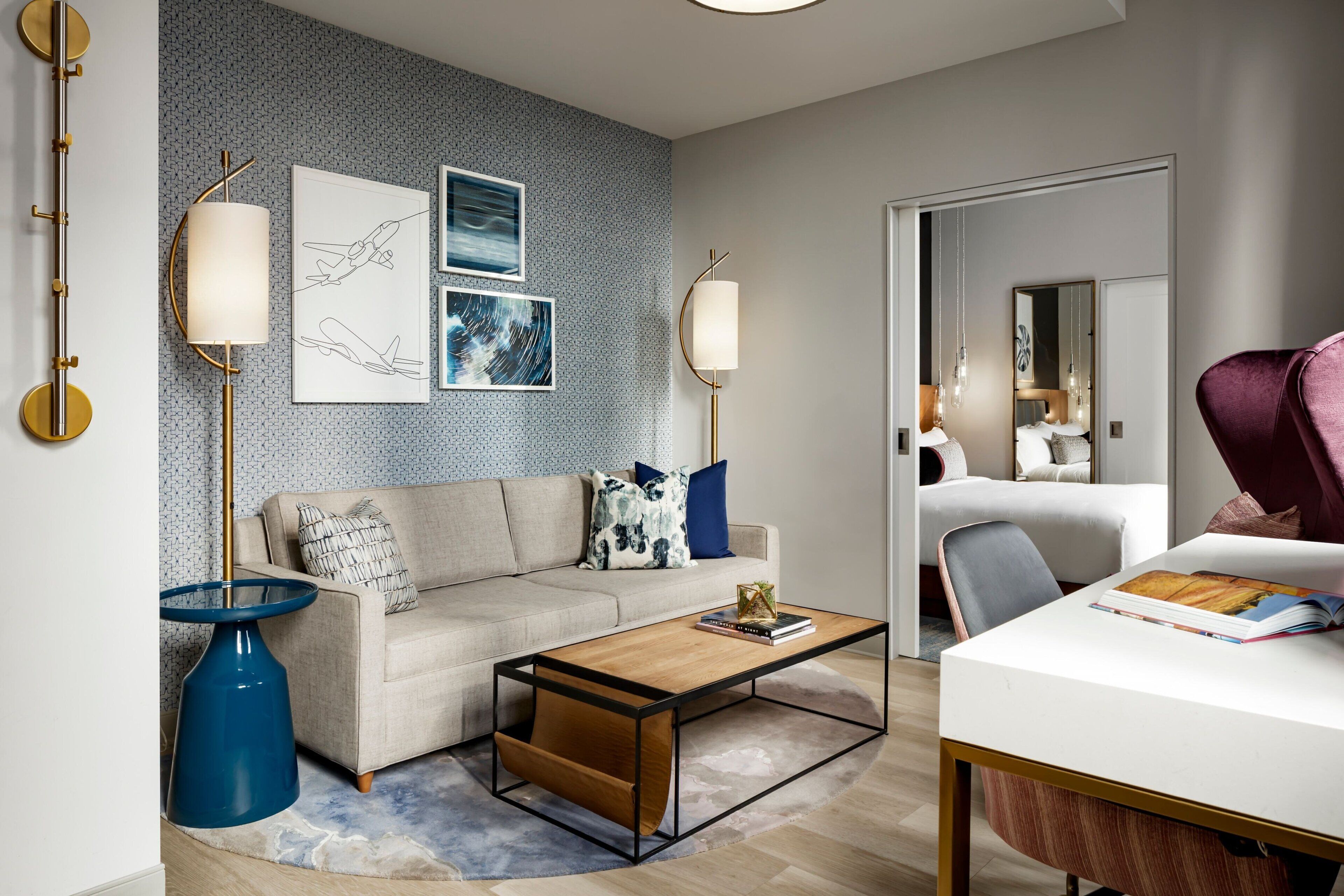 A modern hotel room with a sofa, coffee table, and a desk with a chair. There are wall art pieces and a large mirror.