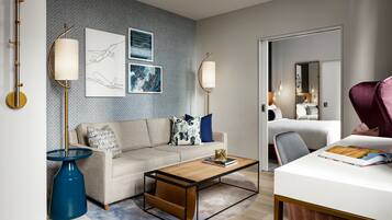 A modern hotel room with a sofa, coffee table, and a desk with a chair. There are wall art pieces and a large mirror.