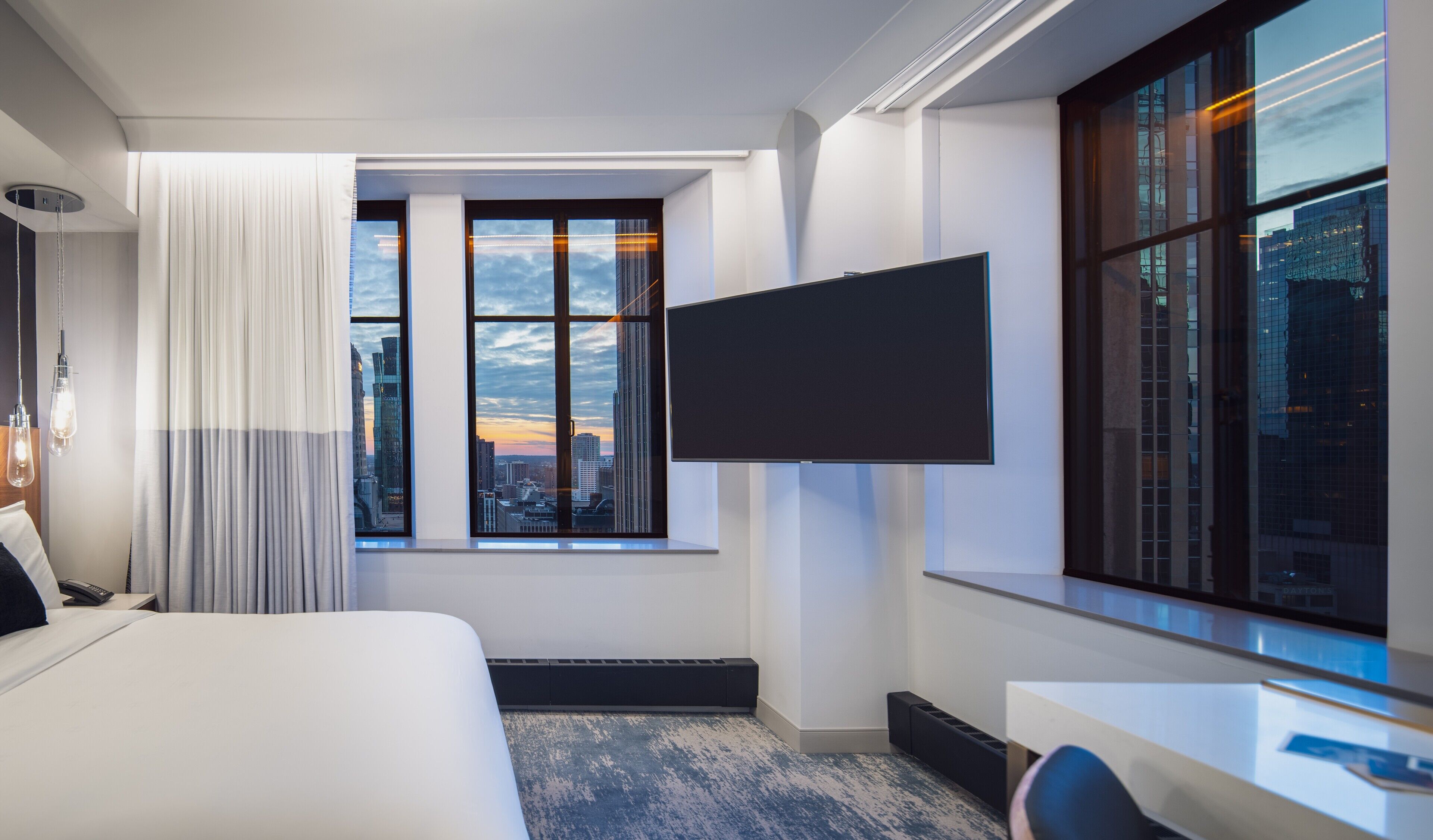 A modern hotel room with a large bed, a flat-screen TV mounted on the wall, a large window with a city view, and a desk with a chair.