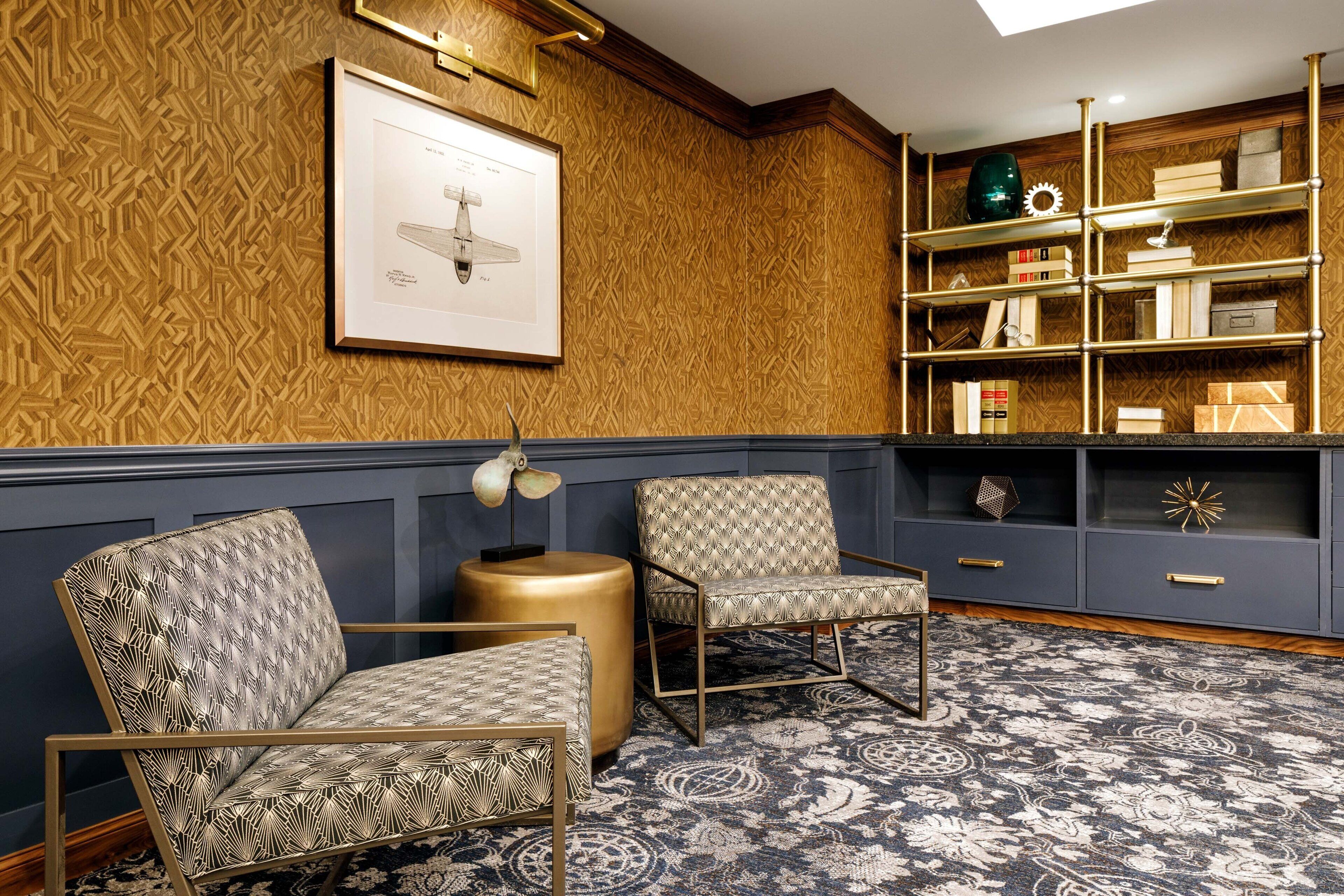 A room with patterned wallpaper, two upholstered chairs, a round gold side table, a blue patterned rug, and a bookshelf with various items.