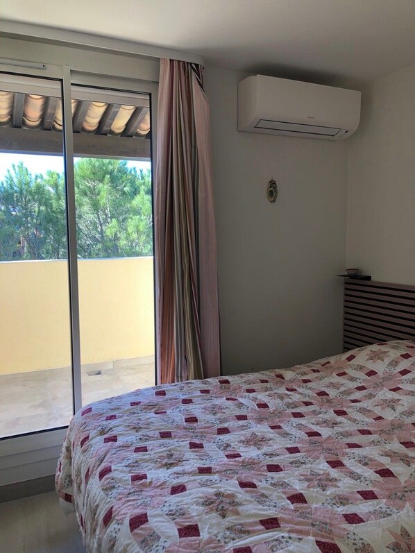 A bedroom with a bed, an air conditioning unit, a window with curtains, and a view of greenery outside.