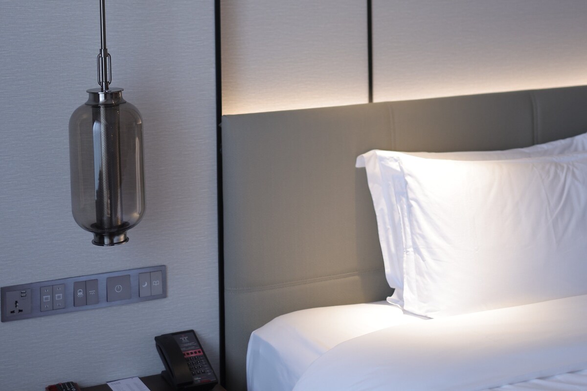 A hotel room with a bed, a wall-mounted light fixture, a control panel with buttons, and a remote control on a bedside table.