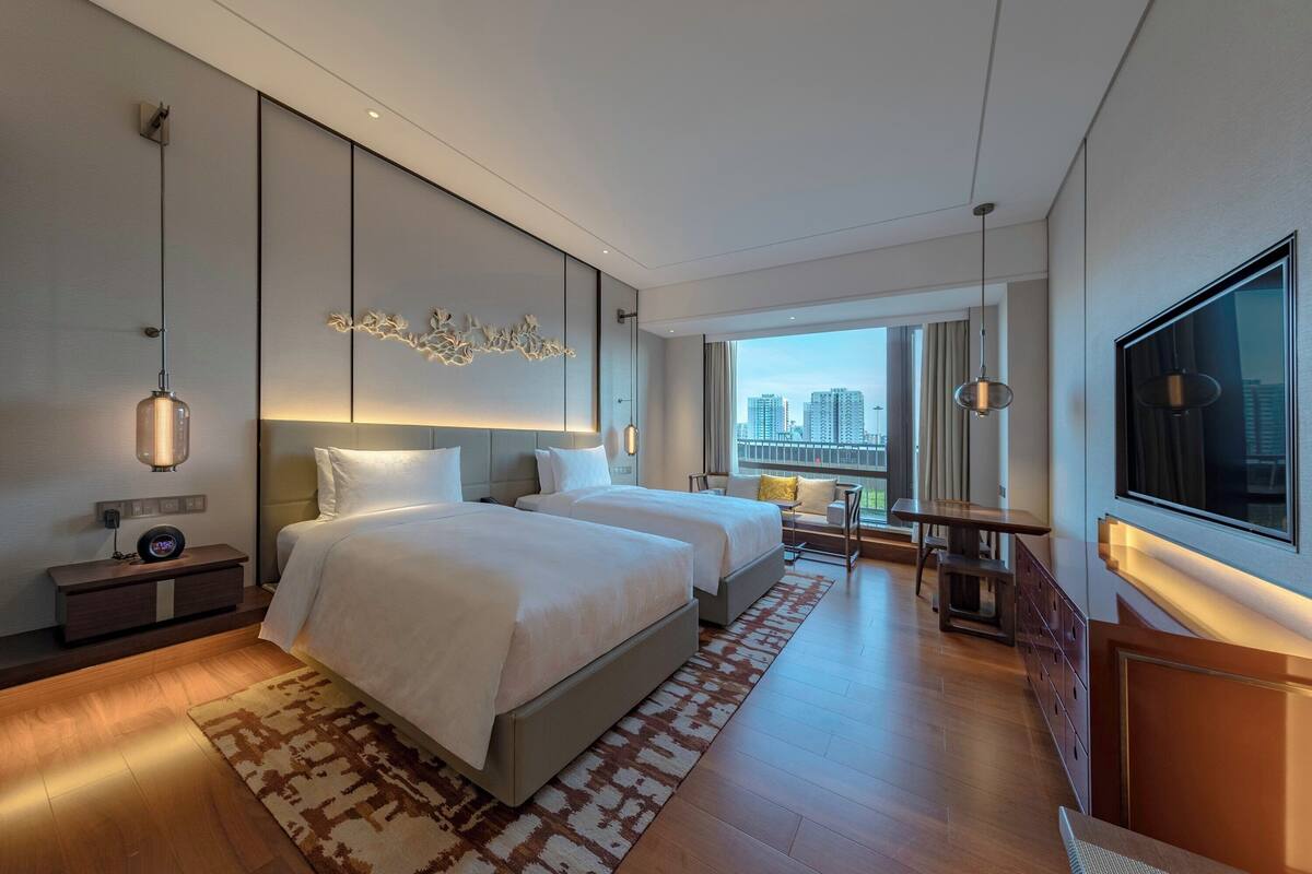 Een moderne hotelkamer met twee bedden, een bureau en een groot raam met uitzicht op de stad.